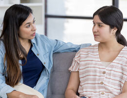 Two women sitting on a couch talking to each other.