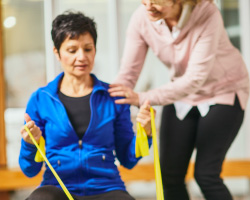 A woman on a balance ball uses a stretch band as a physical therapist stands by.