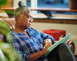 An older woman sits on a couch and writes in a notebook